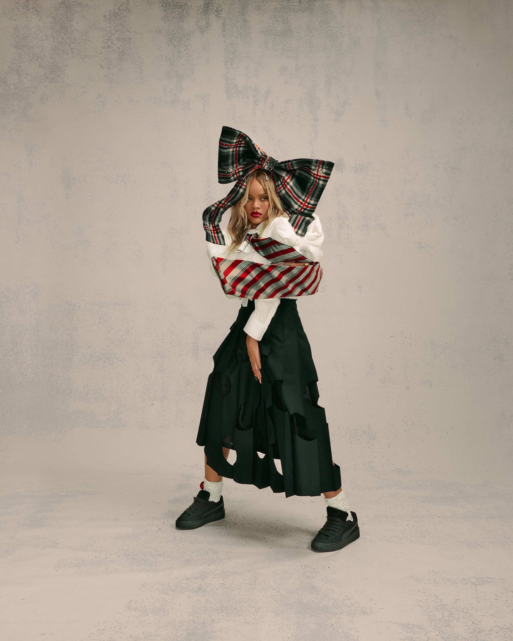 A fashionable young adult female model poses indoors wearing a white top, plaid bow, black skirt, and platform sneakers against a neutral backdrop.