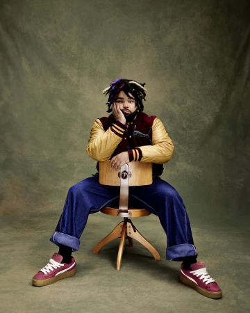 Young man with dreadlocks in a maroon and gold bomber jacket sits on a stool in a studio with a muted green backdrop.