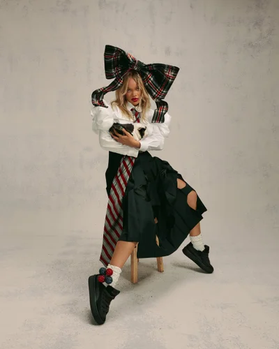 Rihanna seated on a wooden stool wearing a large plaid bow, white blouse, and black sneakers for Fenty x Puma