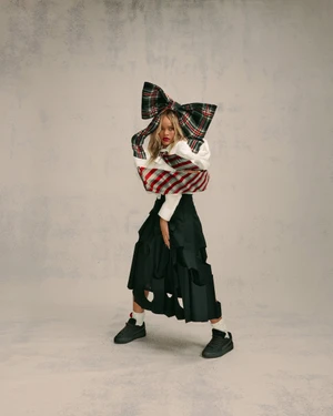 Rihanna wearing an oversized plaid bow and black skirt with sneakers against a grey textured studio backdrop