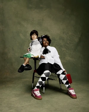 Two models sitting on a vintage school desk wearing white shirts and sneakers against a textured green studio backdrop.