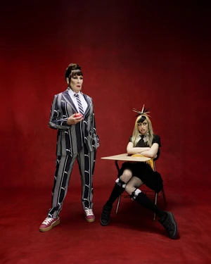 Anjelica Huston standing in a striped suit and Chloe Cherry seated at a school desk against a dark red background for Fenty x Puma