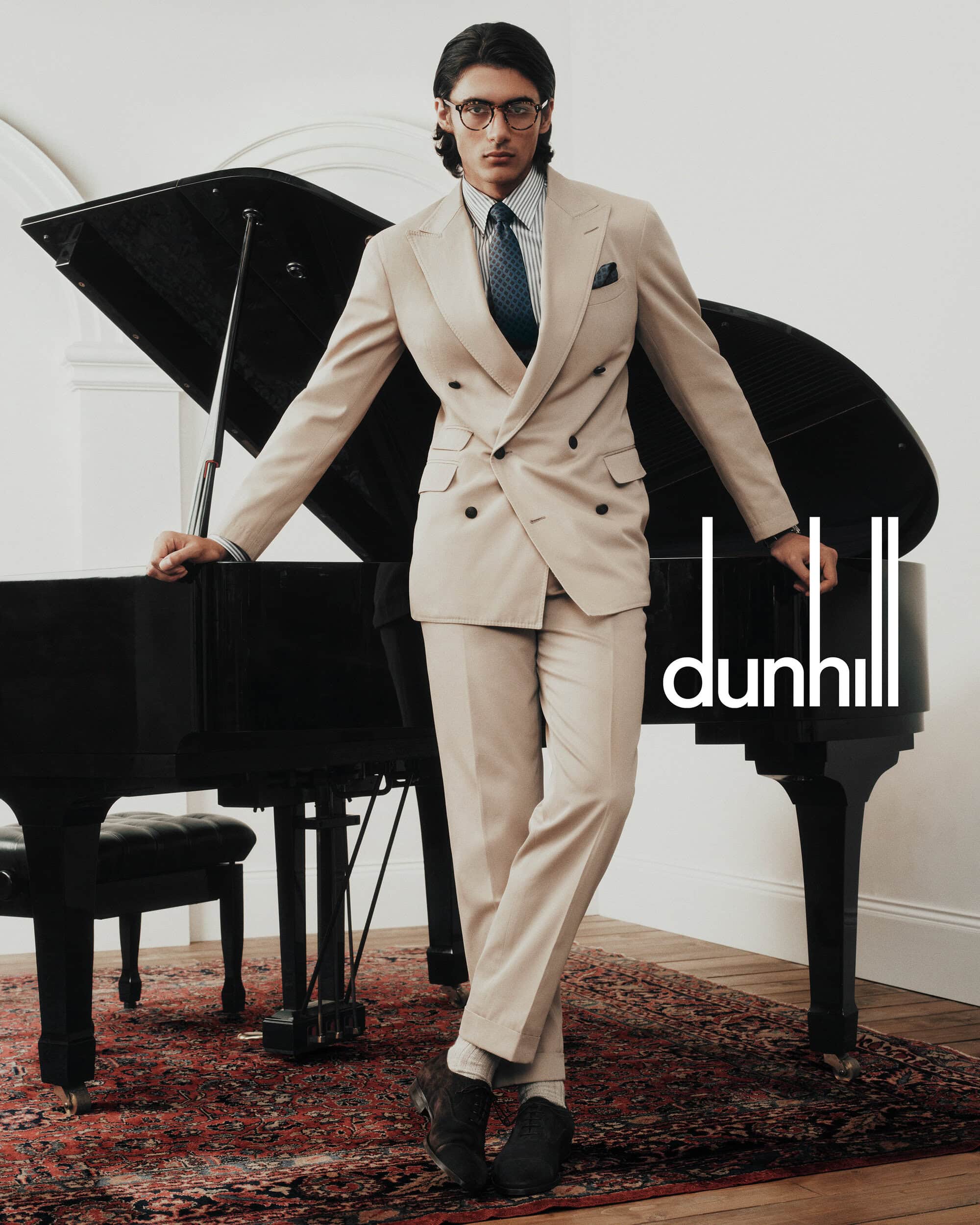Man in light tan suit stands next to piano. The man wears glasses, shirt, and tie. Dunhill brand logo is visible on the right.