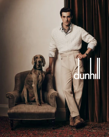 A man leans against a wall next to a dog sitting on an armchair, indoor setting, with Dunhill logo visible in a studio shot.