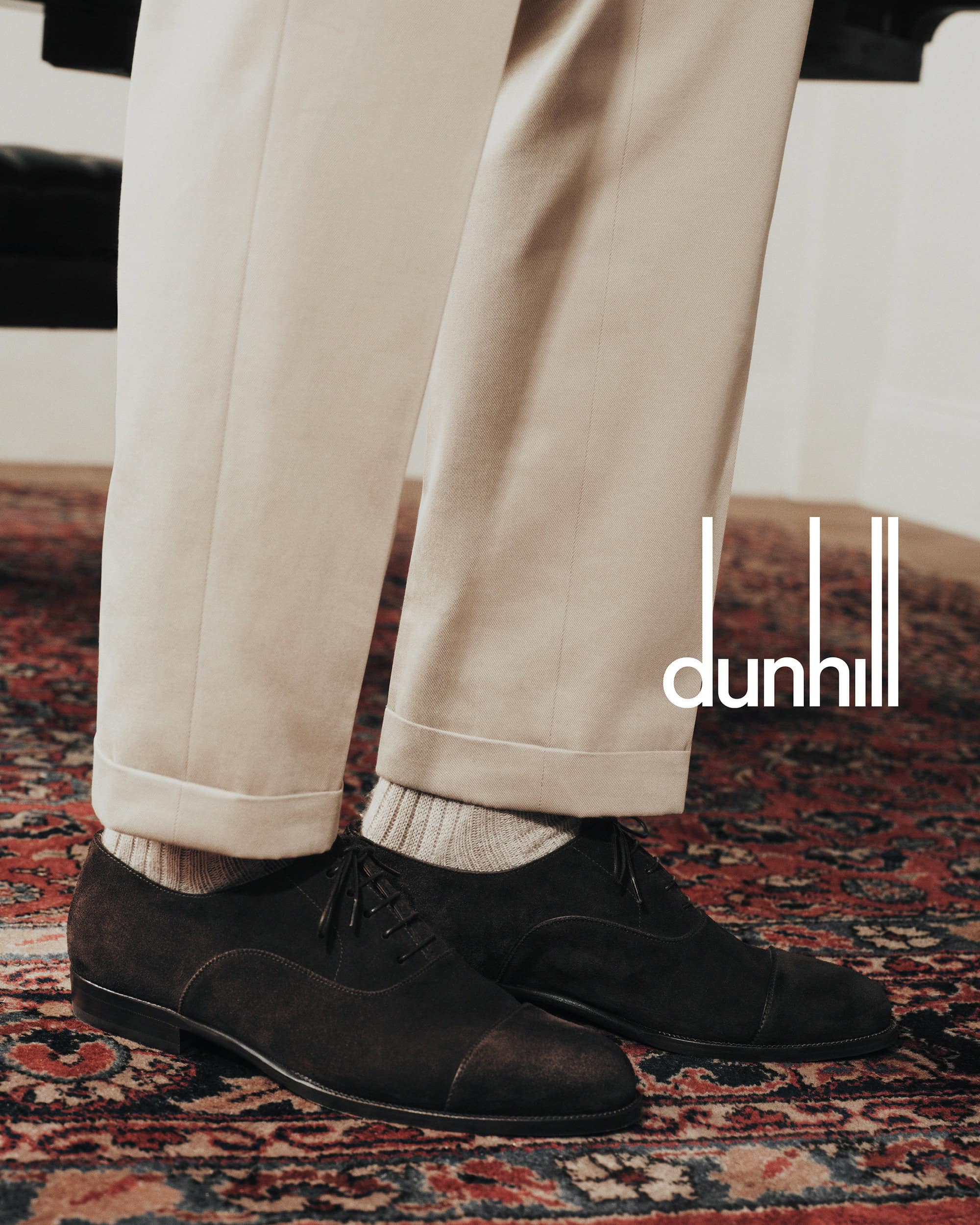 Elevated view of dark brown leather dress shoes with laces, beige socks and pants, set against an ornate rug and background with dunhill logo.