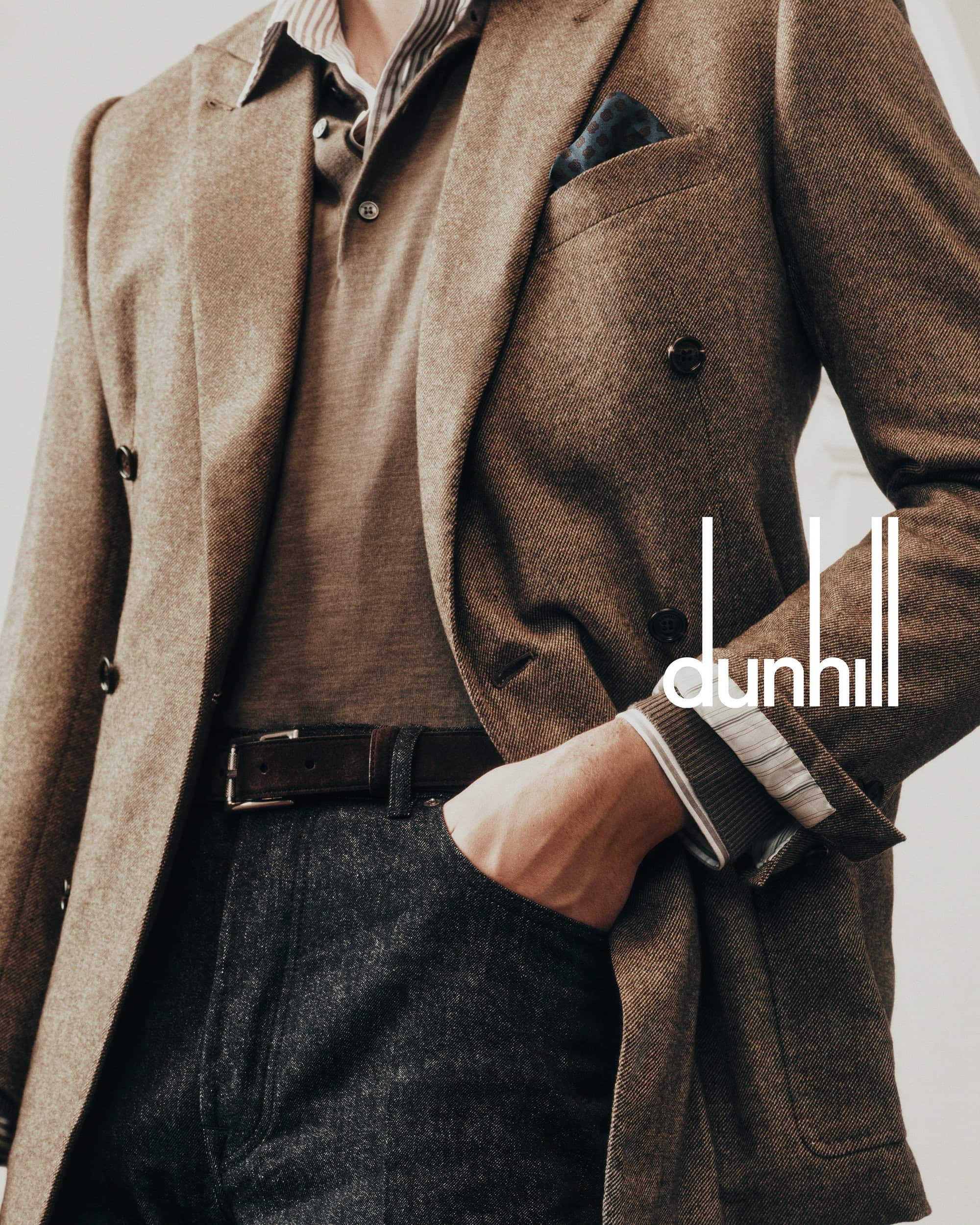 A fashionable adult male is wearing a brown blazer, polo shirt, dark-colored denim jeans, and a belt, posing in a studio with a luxury brand logo.
