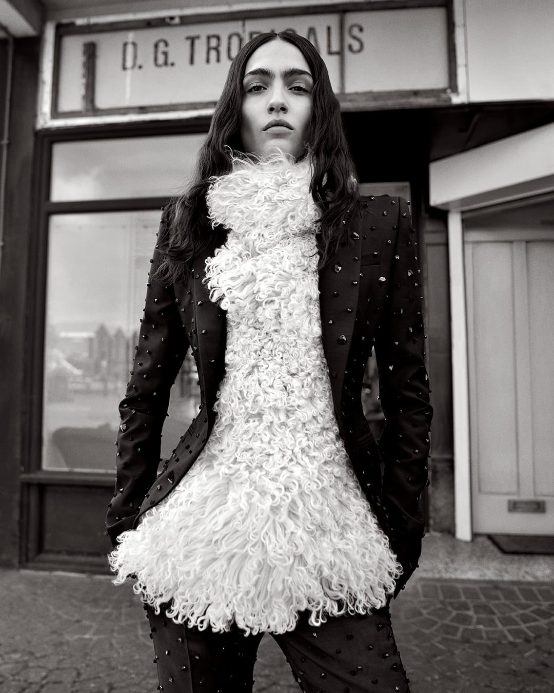 Black and white portrait of a person in a patterned suit and textured white top standing outside a shop with a storefront.