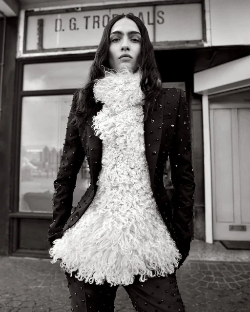 Black and white portrait of a person in a patterned suit and textured white top standing outside a shop with a storefront.