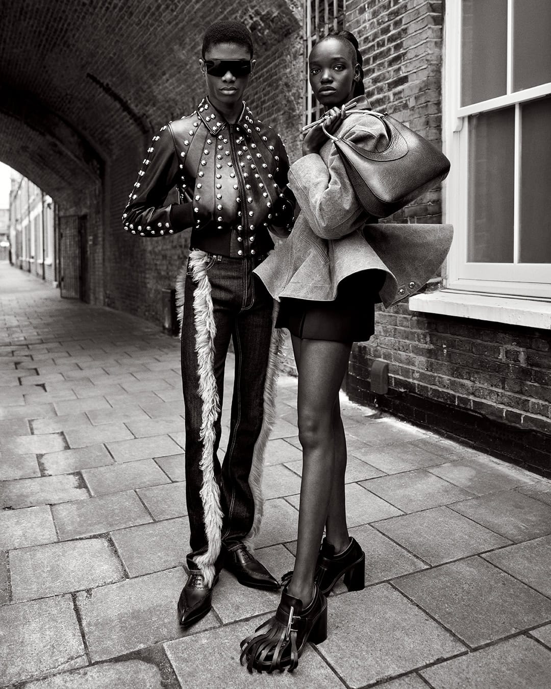 Two Black fashion models pose in a monochromatic outdoor shot wearing stylish clothing: one in a studded leather jacket and the other with a handbag.