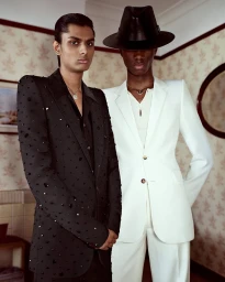 Two male models in tailored suits standing in a room with patterned wallpaper, one in black embellished jacket and one in white suit with hat.