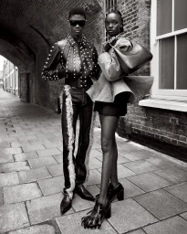 Black and white portrait of two models standing on a paved street wearing studded leather jacket and structured wool coat for McQueen.