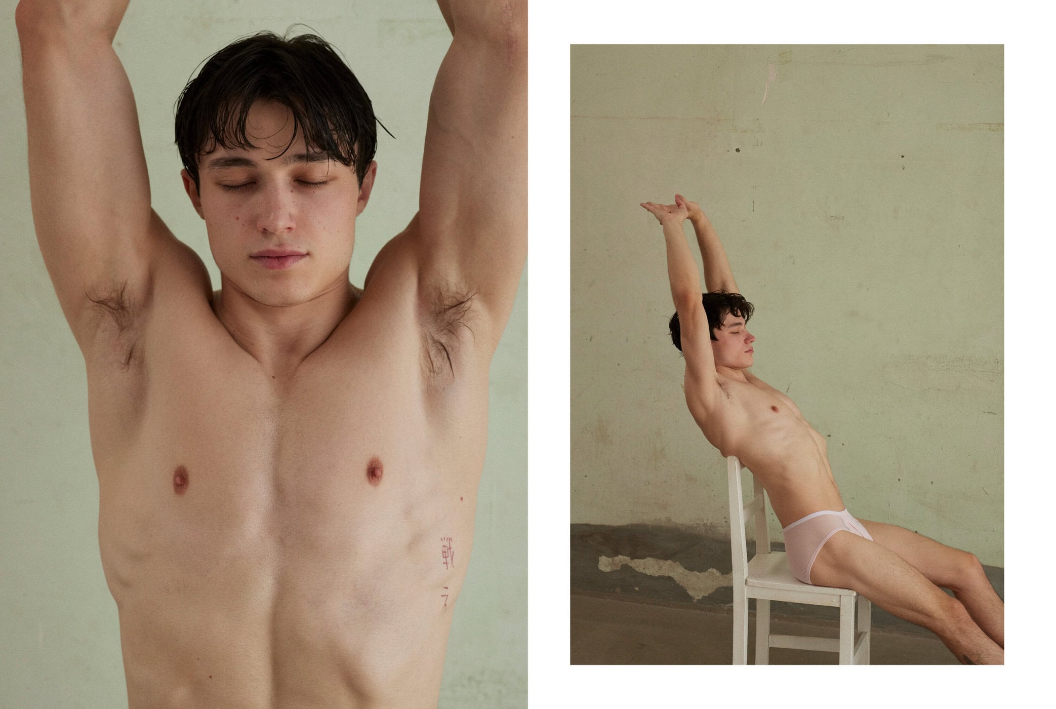 A man with dark hair sits on a white chair, eyes closed, arms stretched overhead, wearing light pink briefs against a plain wall.