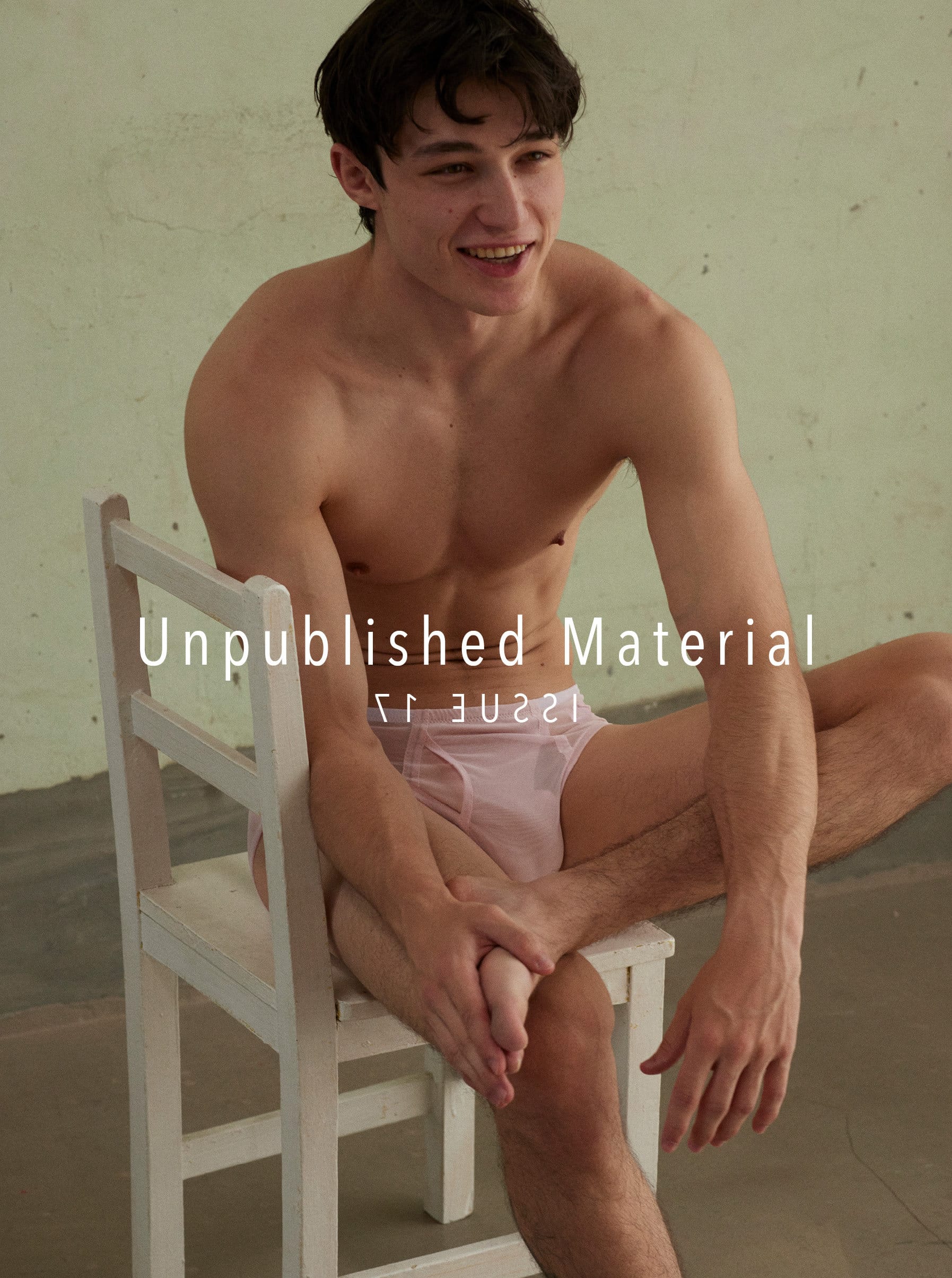 Nude man in briefs sits sideways on a chair looking to the side with a smile in an indoor setting with green wall.