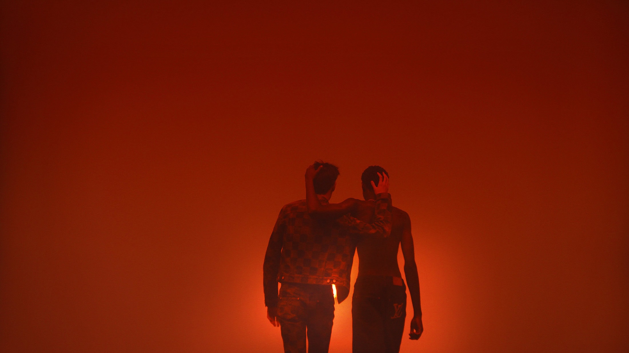 Two people pose in a studio with orange lighting, wearing jackets and pants in a medium shot, creating a dramatic and fashionable silhouette.