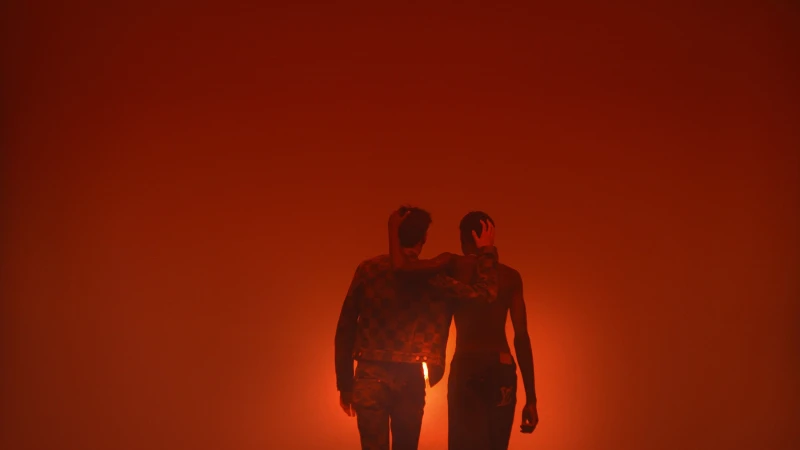 Two people pose in a studio with orange lighting, wearing jackets and pants in a medium shot, creating a dramatic and fashionable silhouette.