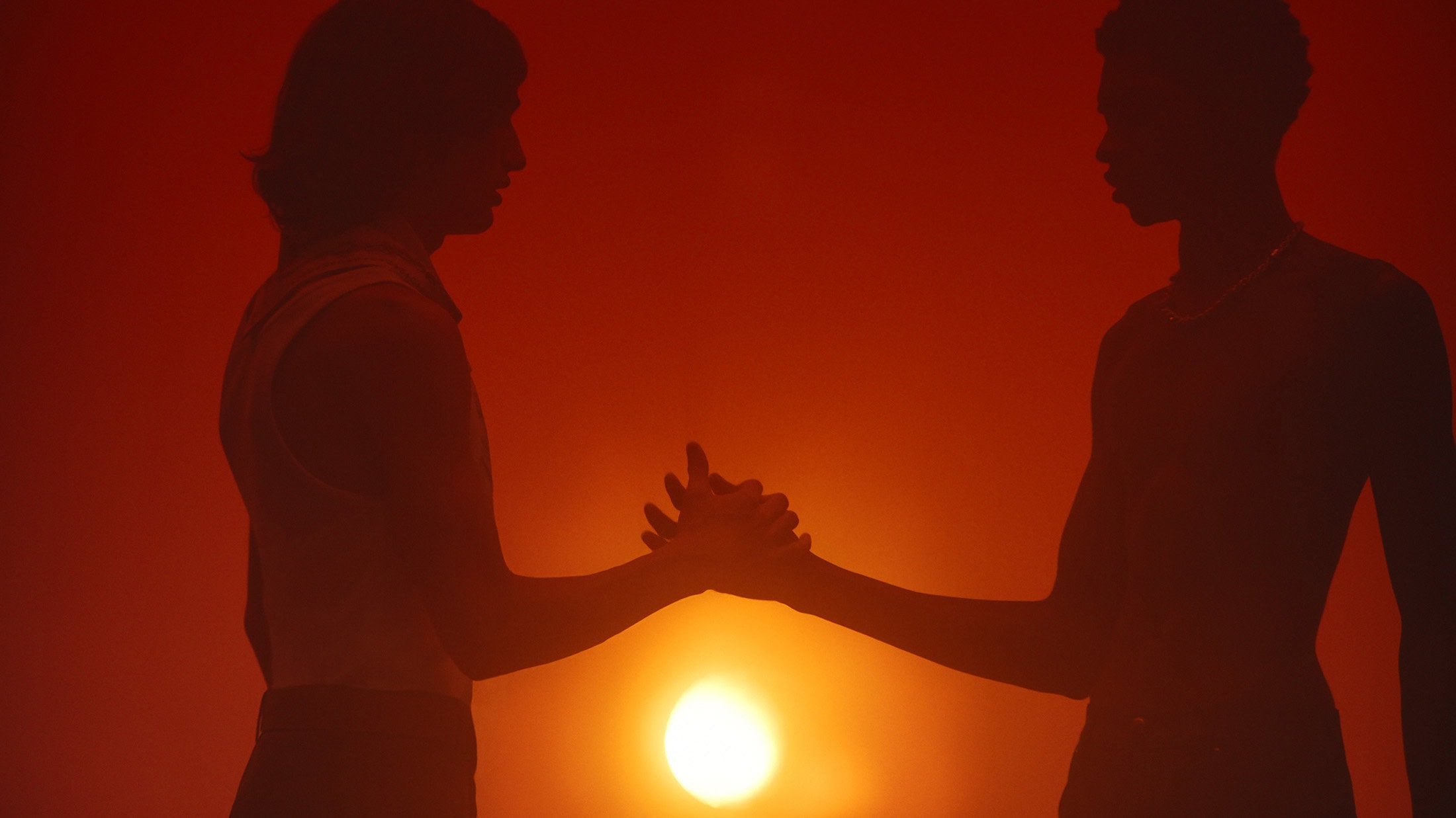 Silhouette of two adults clasping hands against an orange-red sky with a sun-like glow centered between them, creating a dramatic and intimate scene.
