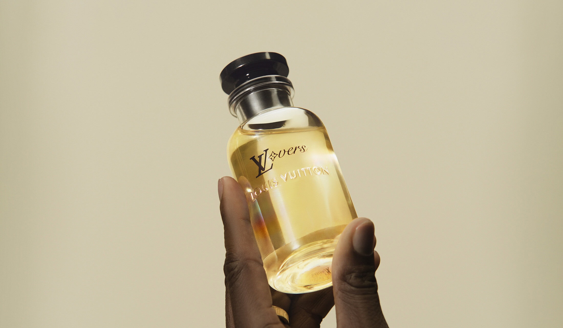 Hand holds glass Louis Vuitton fragrance bottle with gold logo against beige backdrop in medium shot, product-focused beauty still life.