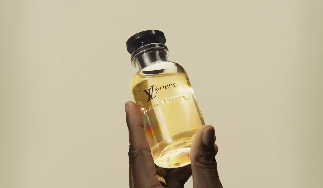 Hand holds glass Louis Vuitton fragrance bottle with gold logo against beige backdrop in medium shot, product-focused beauty still life.