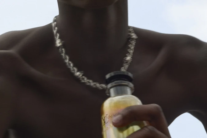 Close-up of a person wearing a twisted silver chain necklace holding a fragrance bottle against an overcast sky