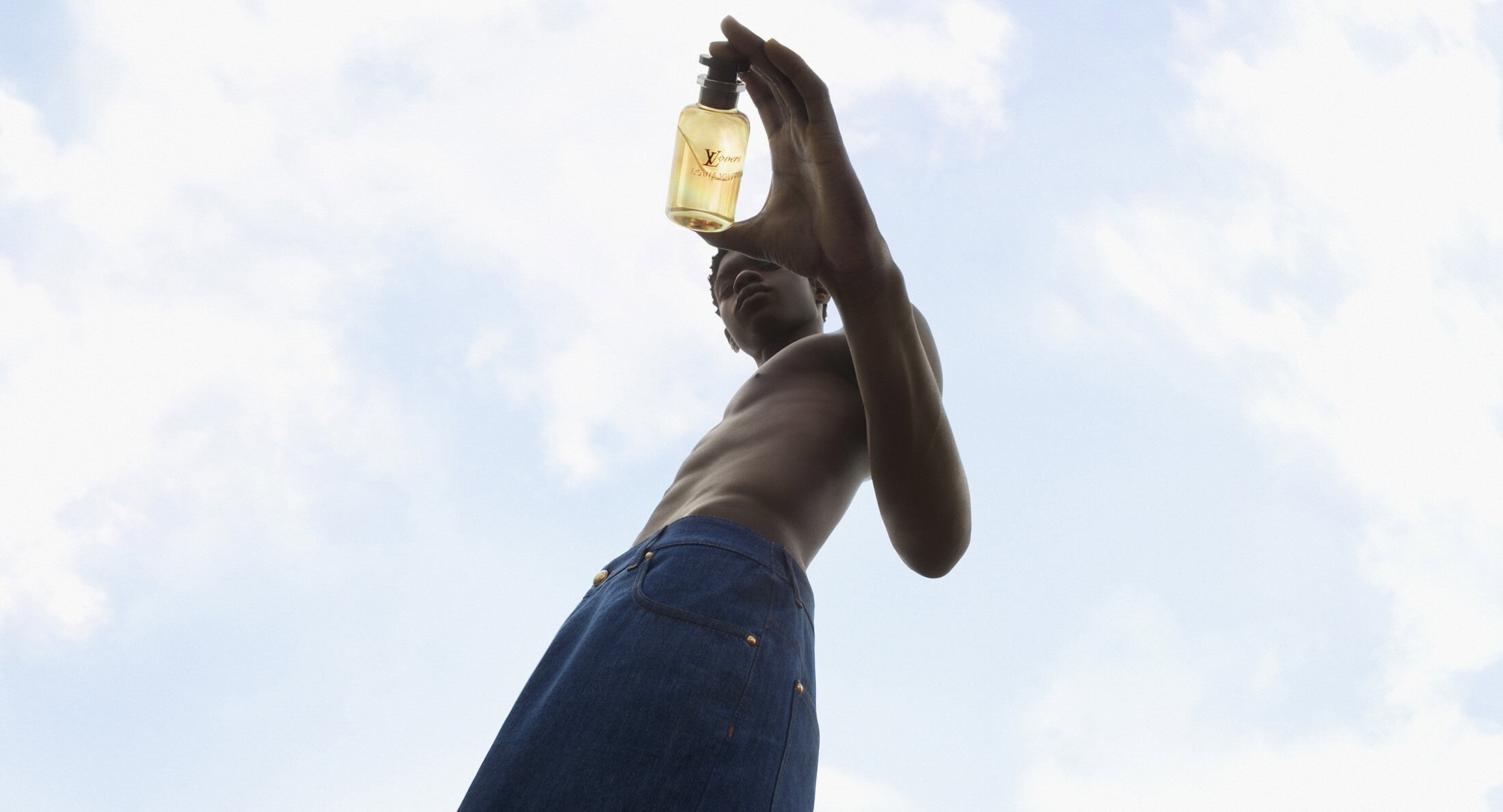 Low angle view of a shirtless Black man holding up a bottle of yellow perfume against a cloudy sky backdrop, shot with natural light.
