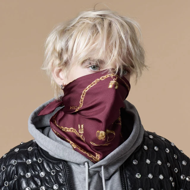 Close-up of person with blonde hair wearing a patterned maroon bandana, grey hoodie, and studded leather jacket against beige backdrop.