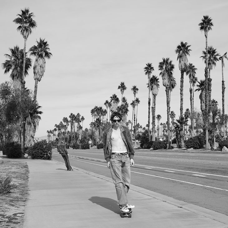 Black and white portrait of a young adult male skateboarding on a sidewalk lined with palm trees and a road with another person in the distance.