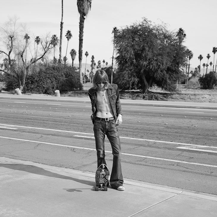 Monochrome portrait of a slender person in a studded jacket and jeans standing with a skateboard in a desert landscape.