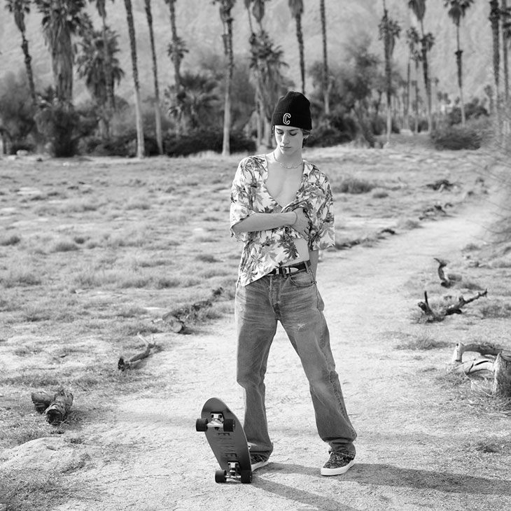 A young adult stands with a skateboard in a monochrome outdoor setting, featuring palm trees and mountains in the backdrop.