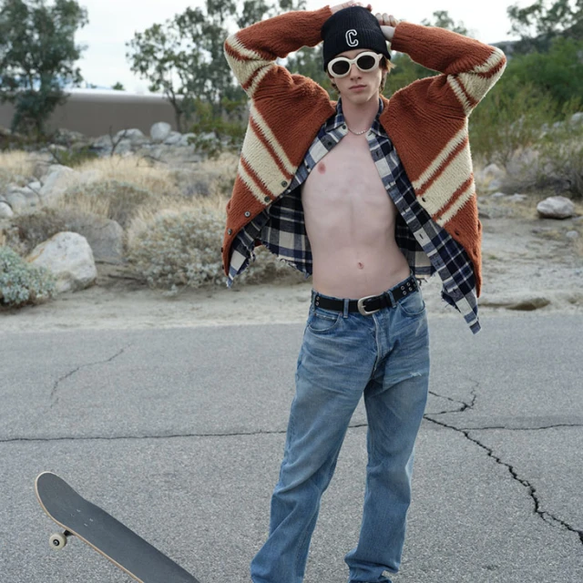 Young man in beanie, sunglasses, cardigan, and jeans standing on road in desert landscape with skateboard. Casual fashion editorial shot.