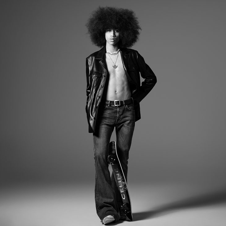 Monochrome portrait of a young male model with a large afro wearing an open leather jacket, jeans, and holding a skateboard.