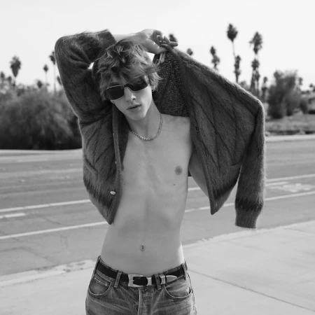 Young man with shaggy hair wearing dark sunglasses and an unbuttoned fuzzy cardigan over jeans standing on a road with palm trees