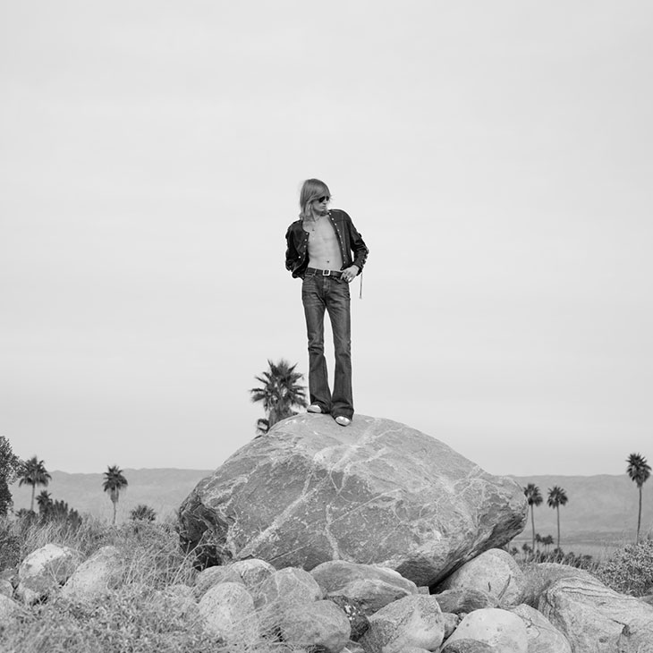 A man with long hair wearing an open leather jacket and flared jeans standing atop a large boulder in a sparse palm tree desert.