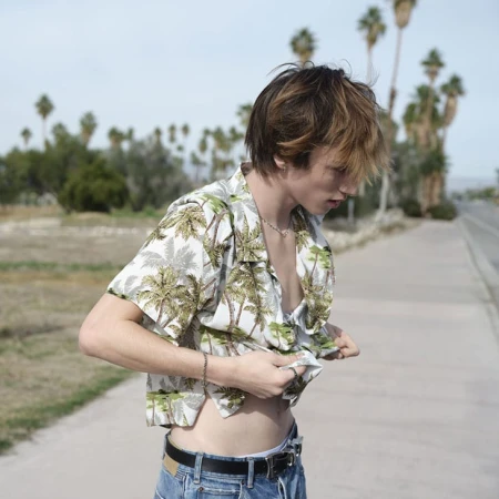 Young adult in Hawaiian shirt poses outdoors on a sunny day with palm trees in background, displaying his torso and wearing jeans.