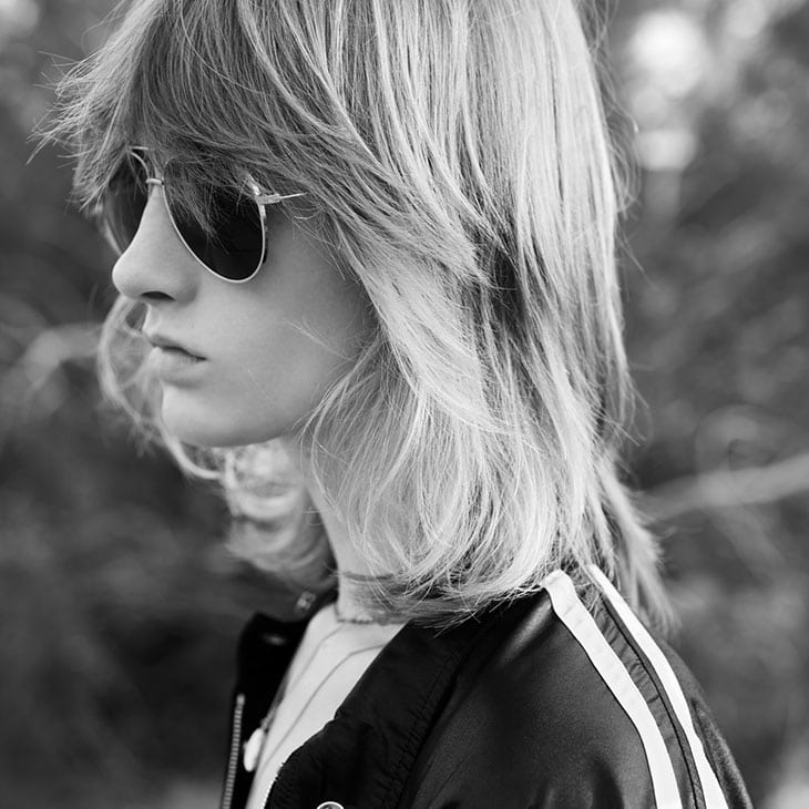 A profile portrait of a young person with long layered blonde hair wearing aviator sunglasses and a track jacket with white stripes.