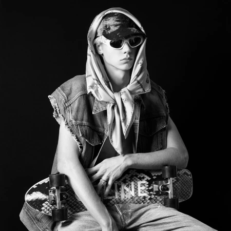 Model in white sunglasses wearing a denim vest and headscarf holding a skateboard against black