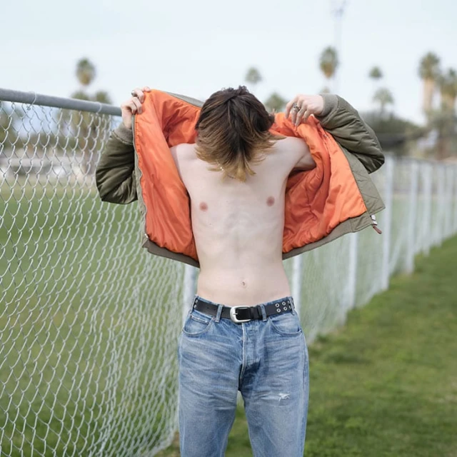 A slender adult stands outdoors, partially undressed. An open olive-green jacket with an orange lining is draped over their shoulders.