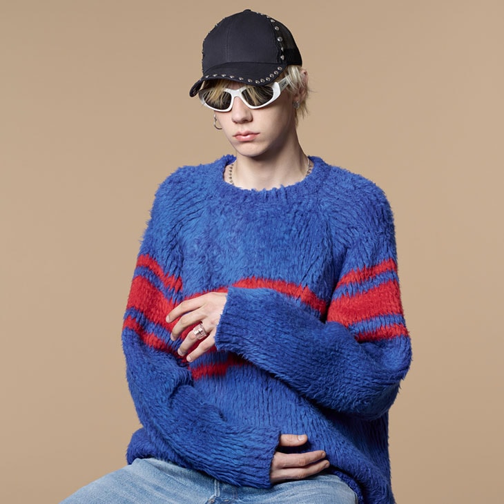 Young adult with blond hair wears a blue and red sweater, cap, and sunglasses against a tan background in a studio portrait.