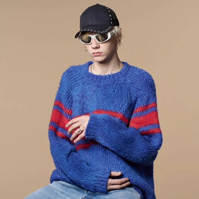 Young adult with blond hair wears a blue and red sweater, cap, and sunglasses against a tan background in a studio portrait.