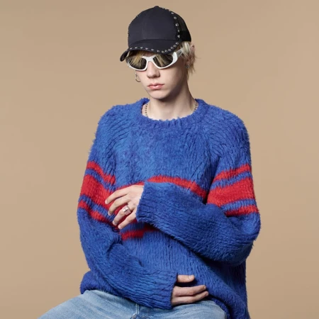 Young adult with blond hair wears a blue and red sweater, cap, and sunglasses against a tan background in a studio portrait.