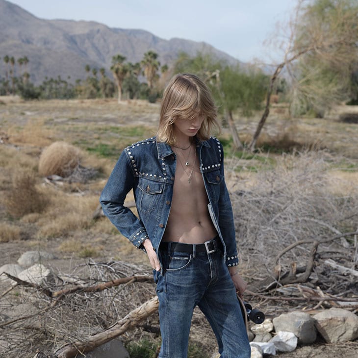 A young adult stands outdoors wearing a denim jacket with studs, denim jeans, and holding a skateboard in a sunny desert landscape.