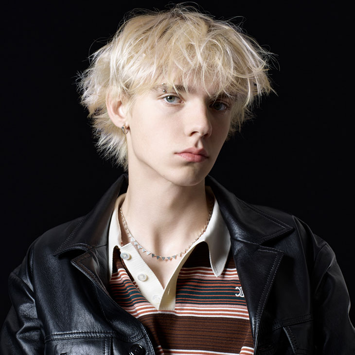 Portrait of a young adult with blond hair, wearing a striped shirt and a black leather jacket against a dark background.