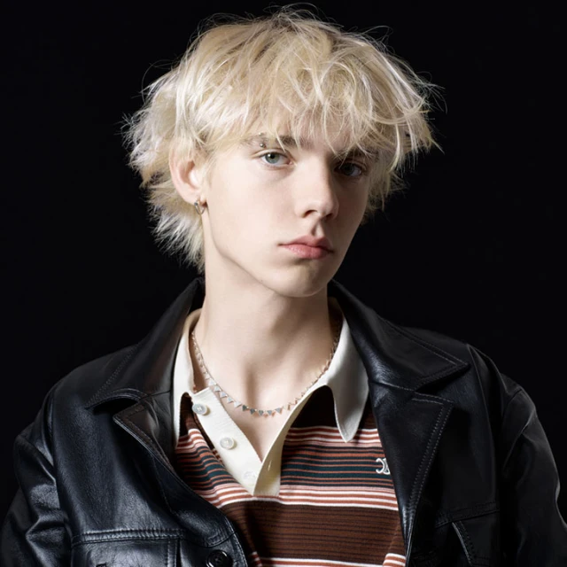 Portrait of a young adult with blond hair, wearing a striped shirt and a black leather jacket against a dark background.