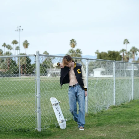 Young man leaning against a chain-link fence outdoors next to a skateboard with loose blue jeans and a varsity jacket.