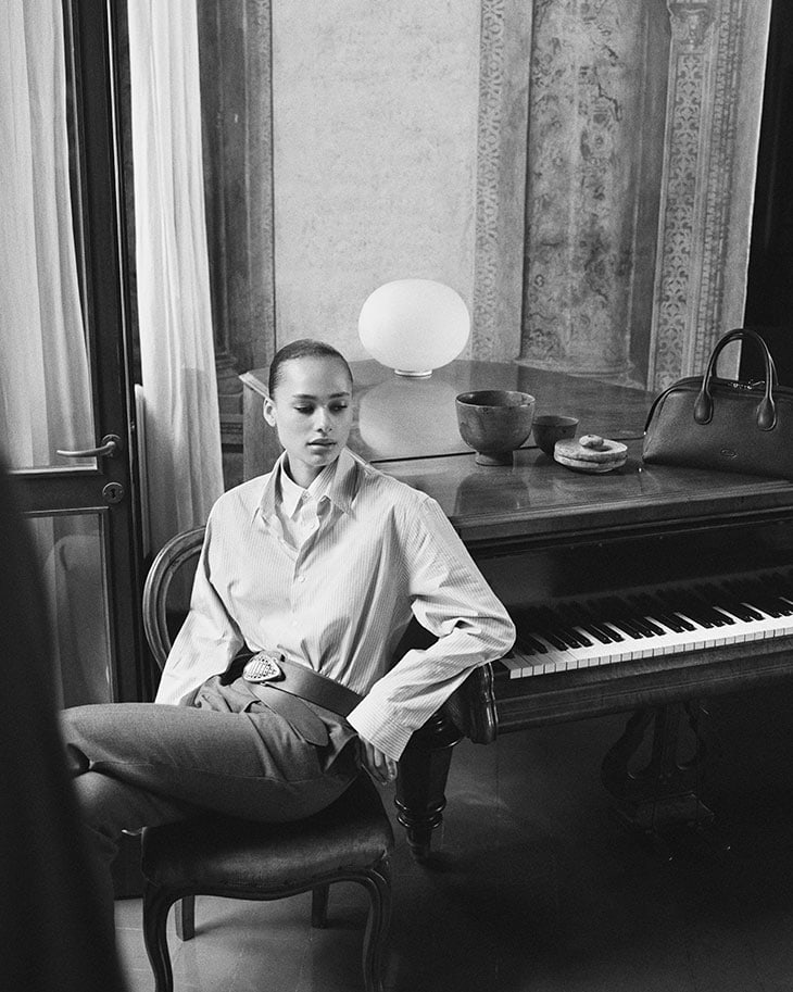 Black and white portrait of a woman seated indoors near a piano wearing a shirt, pants, and a belt, with a lamp and handbag nearby.