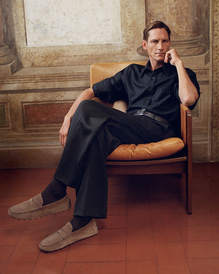 A stylish adult man is sitting in a chair, wearing a dark shirt, pants, and suede loafers in an indoor setting with painted walls.