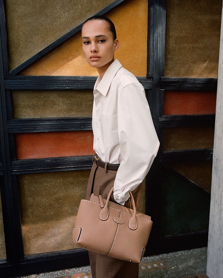 A young woman in a white button-down shirt and brown slacks carries a tan leather handbag in front of a geometric backdrop.