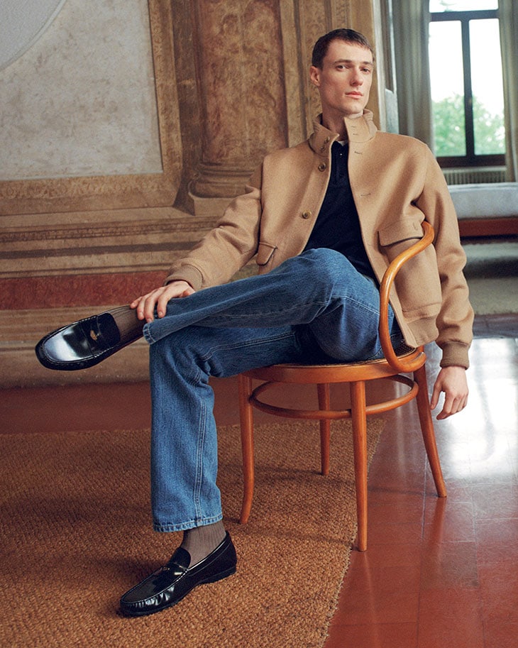 A casually dressed man in a jacket and jeans sits in a chair in an ornate interior, with loafers and neutral colors.