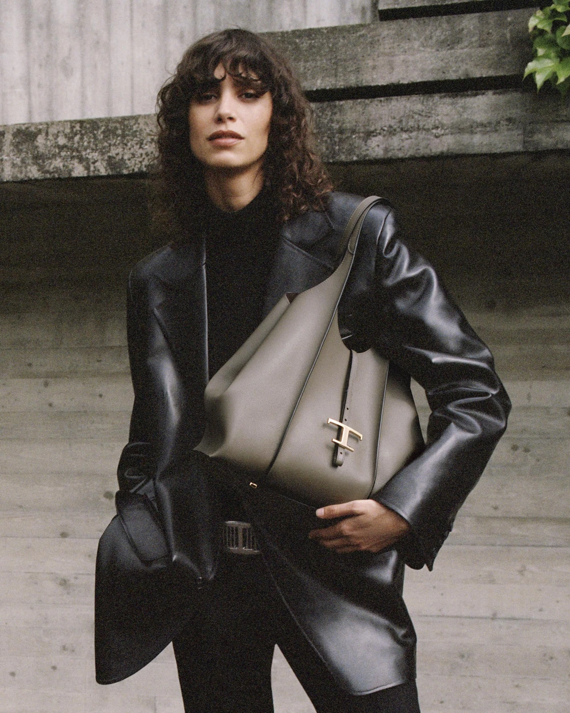 Model in black leather jacket holding a taupe shoulder bag against a concrete background for Tod's Pre-Fall 2024 campaign
