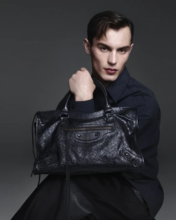 Adult male poses in a photo studio wearing a dark shirt and holding a black textured leather bag. Fashion photography, studio shot.