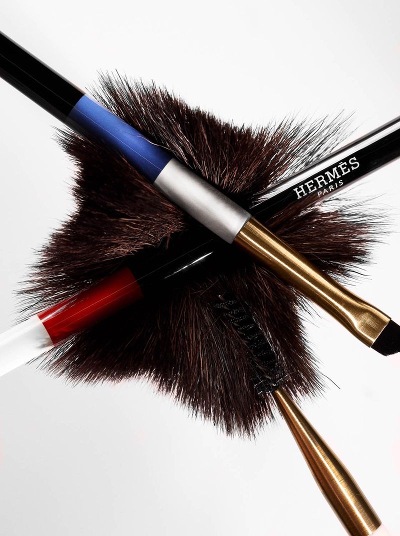 Cosmetic brushes from Hermès Paris sit on a fluffy brown surface against a white background. Focus on beauty and makeup accessories.